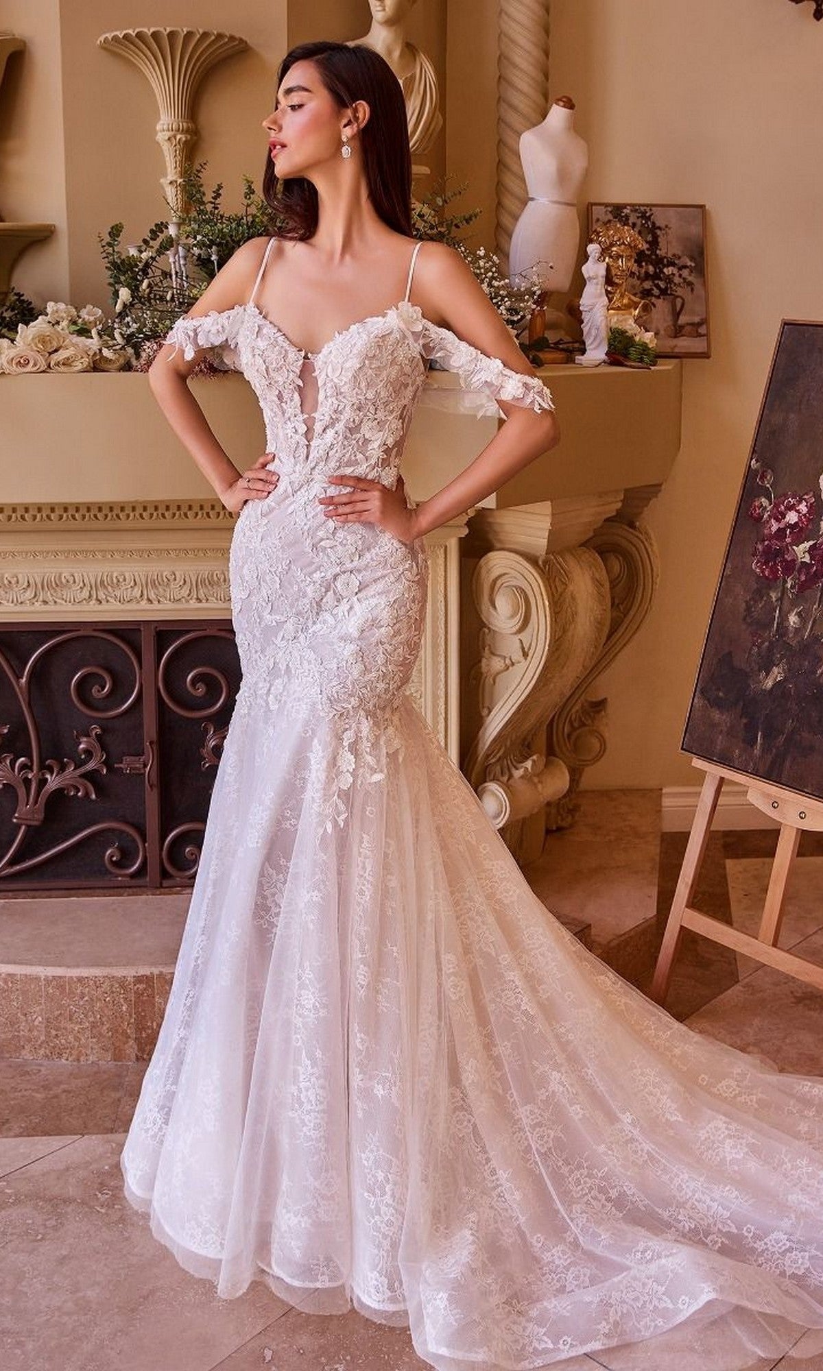A woman stands indoors in the Andrea and Leo WL051 long wedding dress, an off-the-shoulder mermaid gown with lace applique and a long train, surrounded by classical decor and artwork.